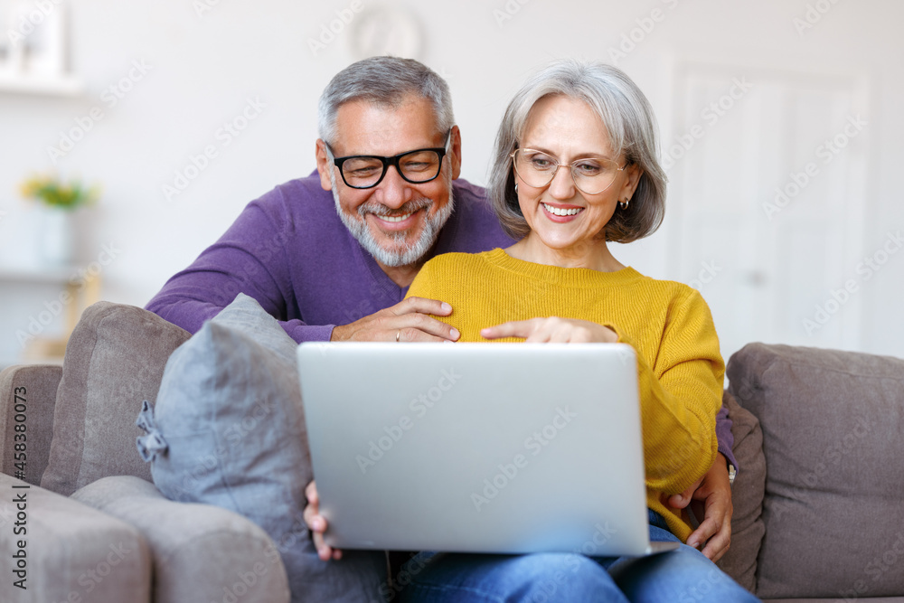 Beautiful mature family couple looking at laptop with smile on face while spending time together Beautiful mature family couple looking at laptop with smile on face while spending time together