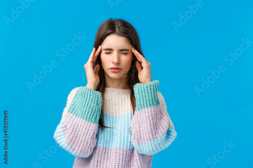 Hungover, health and people concept. Stressed young woman feeling headache, close eyes touching temples, trying easy pain with medicine, suffering migraine, standing blue background photo