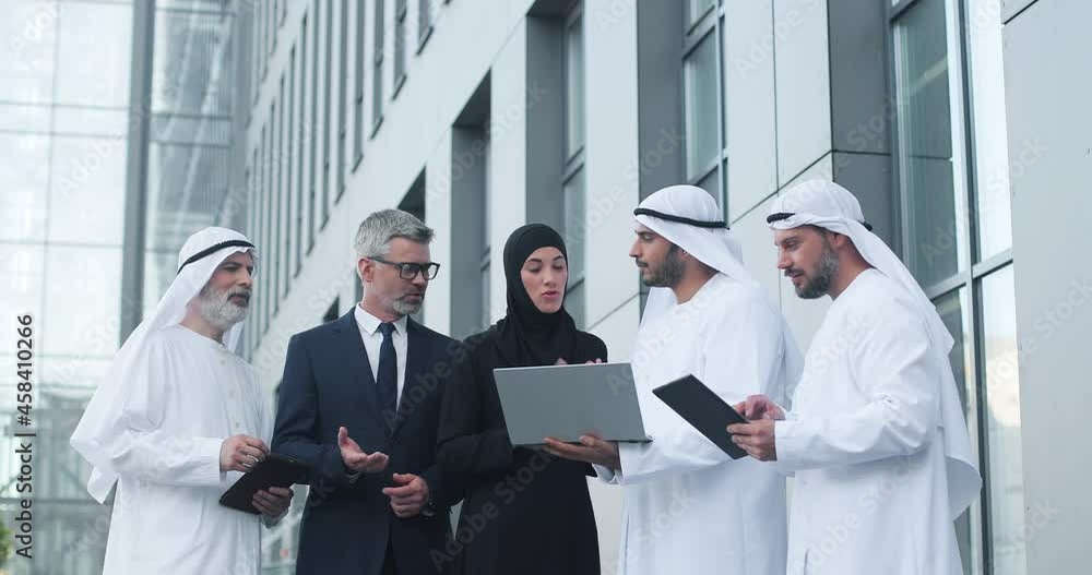 Waist up portrait of young multi ethnic entrepreneurs standing near the modern office at team meeting and speaking. Caucasian man discussing business ideas with muslim colleagues at the conference Waist up portrait of young multi ethnic entrepreneurs standing near the modern office at team meeting and speaking. Caucasian man discussing business ideas with muslim colleagues at the conference