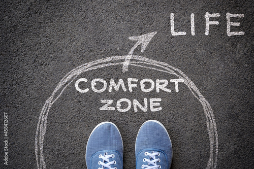 Exit from comfort zone concept. Feet in blue jeans sneakers standing inside circle comfort zone and outward arrow chalky on the asphalt.