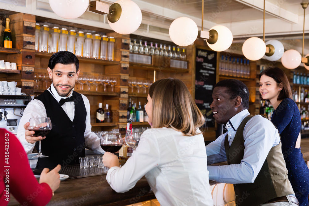 Barman is servicing young cheerful positive people who are relaxing in bar indoor.