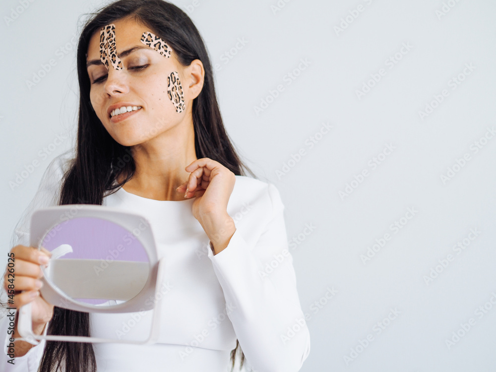 Young beautiful brunette woman with long hair with leopard tapes on her face looking at mirror Young beautiful brunette woman with long hair with leopard tapes on her face looking at mirror