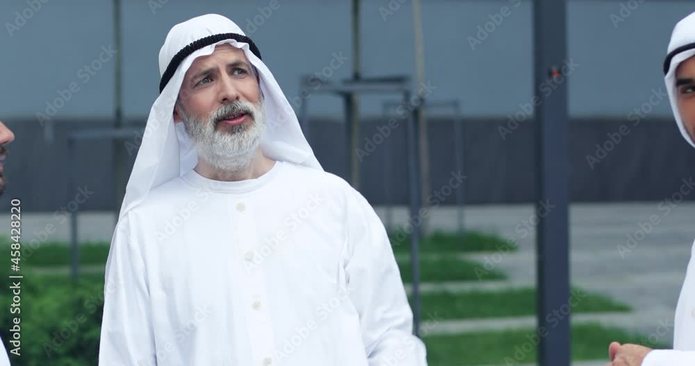 Waist up portrait view of the confident senior sheikh chatting with his business partners and gesturing while standing at the street. Business concept Waist up portrait view of the confident senior sheikh chatting with his business partners and gesturing while standing at the street. Business concept