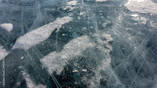 Turquoise transparent ice of a frozen lake. Close-up. Full-screen mode. Deep intersecting cracks, bubbles of frozen methane gas are visible. A little snow on the surface. Baikal