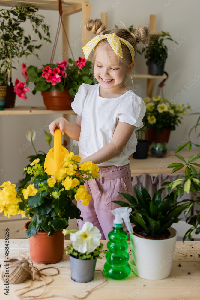 a little blonde girl with a headband on her head and wearing work gloves sprays or waters indoor flowers. Concept of spring time, home gardening, child house-help. Lifestyle, space for text. 