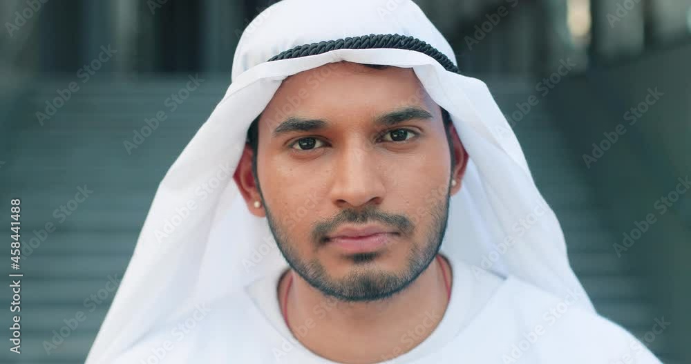 Portrait view of the young arabian man wearing kandura looking at the camera and smiling with pleasure while spending time at the street near his office Portrait view of the young arabian man wearing kandura looking at the camera and smiling with pleasure while spending time at the street near his office