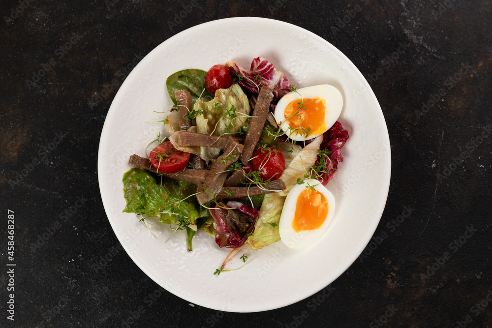 A beautiful Fresh salad with meat tongue vegetables lettuce leaves egg cucumbers bell pepper Cherry tomatoes olives red onions is decorated on a white plate on a black background large serving A beautiful Fresh salad with meat tongue vegetables lettuce leaves egg cucumbers bell pepper Cherry tomatoes olives red onions is decorated on a white plate on a black background large serving