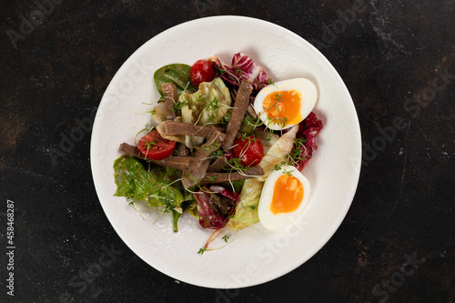 A beautiful Fresh salad with meat tongue vegetables lettuce leaves egg cucumbers bell pepper Cherry tomatoes olives red onions is decorated on a white plate on a black background large serving 