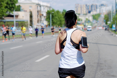 Young woman in sportswear with smartphone attached to her shoulder running city marathon, other contestants running towards her. Concept of sport