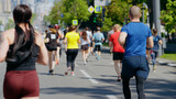 Contestants of city marathon running on asphalt road, focus on woman with ponytail, buildings on blurred background. Back view athletic people taking part in sports competition