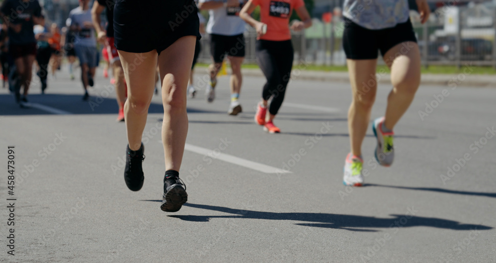 Legs of athletes running marathon on asphalt road, focus on female legs, disabled person riding pumped wheelchair on background. Partial view of sports event contestants