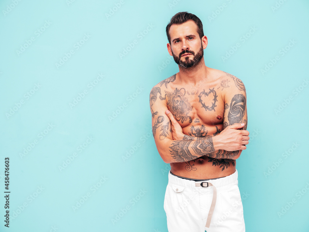 Fototapeta premium Portrait of handsome confident stylish hipster lambersexual model. Sexy modern man. Naked torso with tattoos.Fashion male posing in studio near light blue wall. Crossed arms
