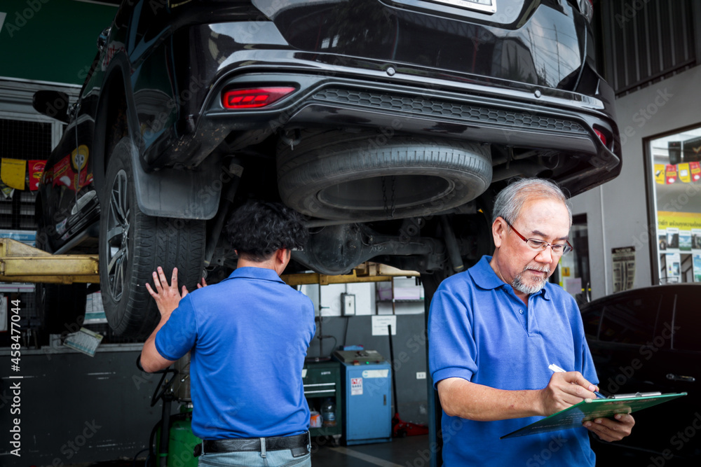 Senior foreman checking list of vehicle repair service while two mechanic checking wheel, inspecting under car body of customer car at automobile garage service, vehicle repair service shop concept
