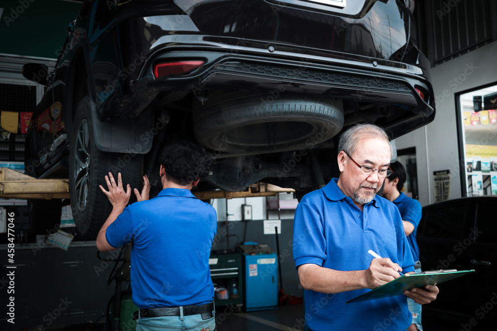 Senior foreman checking list of vehicle repair service while two mechanic checking wheel, inspecting under car body of customer car at automobile garage service, vehicle repair service shop concept Senior foreman checking list of vehicle repair service while two mechanic checking wheel, inspecting under car body of customer car at automobile garage service, vehicle repair service shop concept