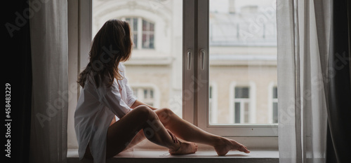 Beautiful sensual young woman with long hair in a white shirt elegantly sitting on the windowsill in an apartment with city view