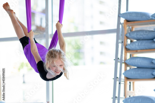  young smiling girl practice in  stretching swing in purple hammock in fitness club. kids Aerial flying yoga exercises.