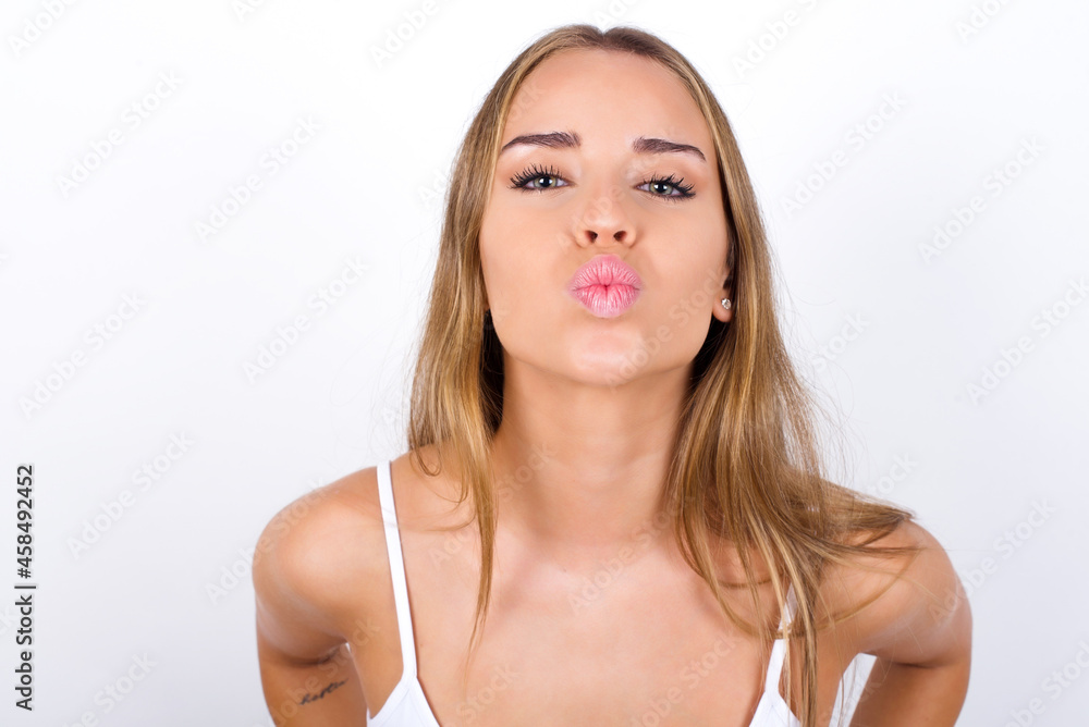 beautiful blonde girl wearing sexy t-shirt on white background, keeps lips as going to kiss someone, has glad expression, grimace face. Standing indoors. Beauty concept.