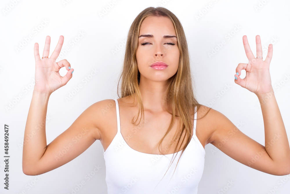 beautiful blonde girl wearing sexy t-shirt on white background doing yoga, keeping eyes closed, holding fingers in mudra gesture. Meditation, religion and spiritual practices. beautiful blonde girl wearing sexy t-shirt on white background doing yoga, keeping eyes closed, holding fingers in mudra gesture. Meditation, religion and spiritual practices.