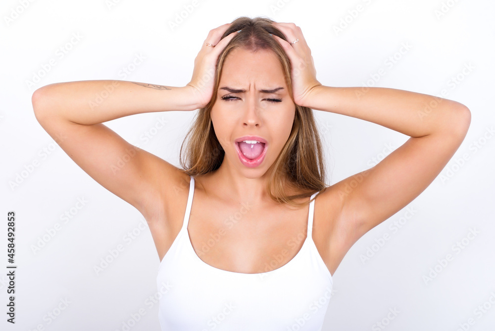 Fototapeta premium Shocked panic beautiful blonde girl wearing sexy t-shirt on white background holding hands on head and screaming in despair and frustration.