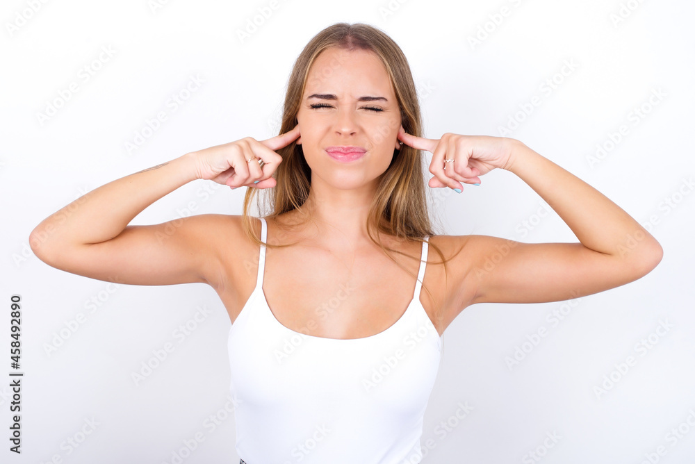 Stop making this annoying sound! Unhappy stressed out beautiful blonde girl wearing sexy t-shirt on white background making worry face, plugging ears with fingers, irritated with loud noise. Stop making this annoying sound! Unhappy stressed out beautiful blonde girl wearing sexy t-shirt on white background making worry face, plugging ears with fingers, irritated with loud noise.