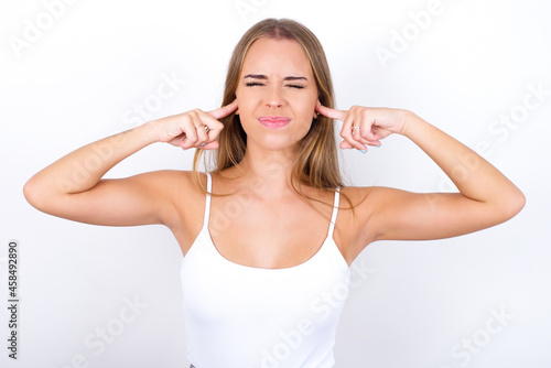 Stop making this annoying sound! Unhappy stressed out beautiful blonde girl wearing sexy t-shirt on white background making worry face, plugging ears with fingers, irritated with loud noise.