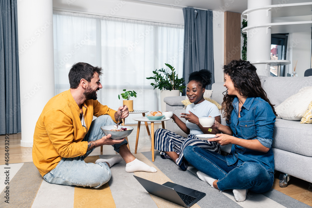 Three young carefree friends two women and one man sitting in the apartment on the floor listening to music on a laptop eating and having fun for a free weekend without worries Three young carefree friends two women and one man sitting in the apartment on the floor listening to music on a laptop eating and having fun for a free weekend without worries