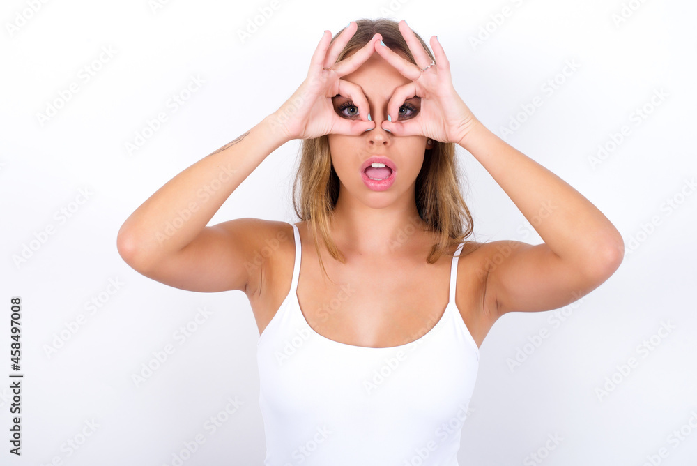 Playful excited beautiful blonde girl wearing sexy t-shirt on white background showing Ok sign with both hands on eyes, pretending to wear spectacles.