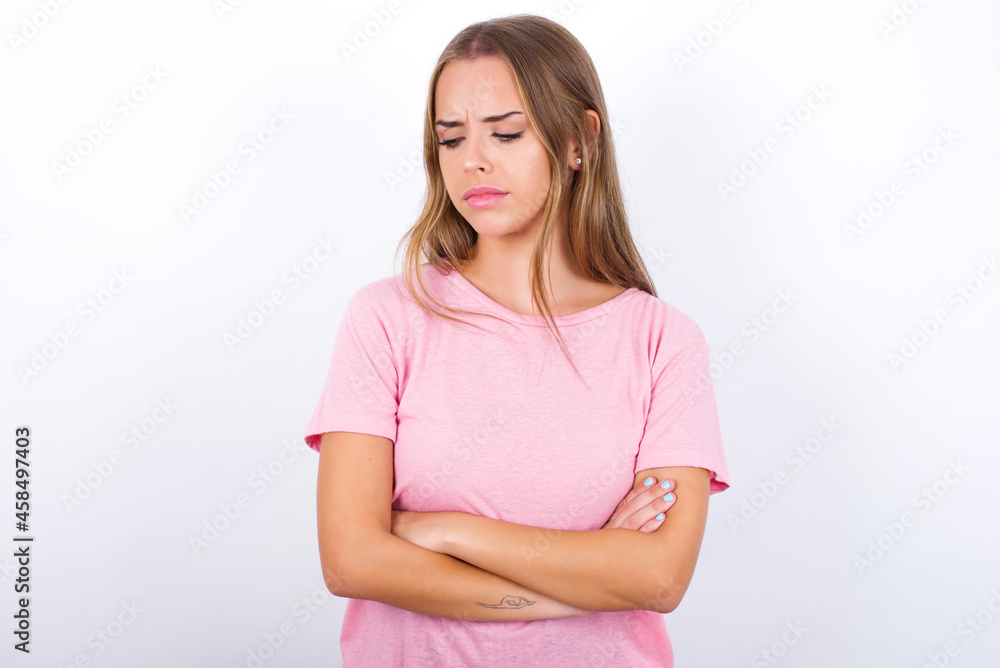Displeased beautiful blonde girl wearing pink t-shirt on white background with bad attitude, arms crossed looking sideways. Negative human emotion facial expression feelings. Displeased beautiful blonde girl wearing pink t-shirt on white background with bad attitude, arms crossed looking sideways. Negative human emotion facial expression feelings.