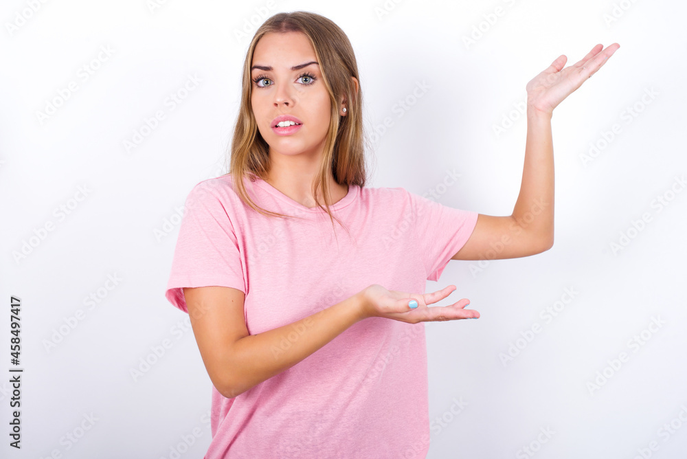 beautiful blonde girl wearing pink t-shirt on white background pointing aside with both hands showing something strange and saying: I don't know what is this. Advertisement concept. beautiful blonde girl wearing pink t-shirt on white background pointing aside with both hands showing something strange and saying: I don't know what is this. Advertisement concept.