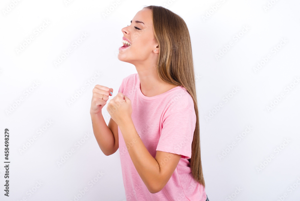 beautiful blonde girl wearing pink t-shirt on white background excited and glad to achieve victory, clenches fists, screams in excitement with closed eyes,successful person. beautiful blonde girl wearing pink t-shirt on white background excited and glad to achieve victory, clenches fists, screams in excitement with closed eyes,successful person.