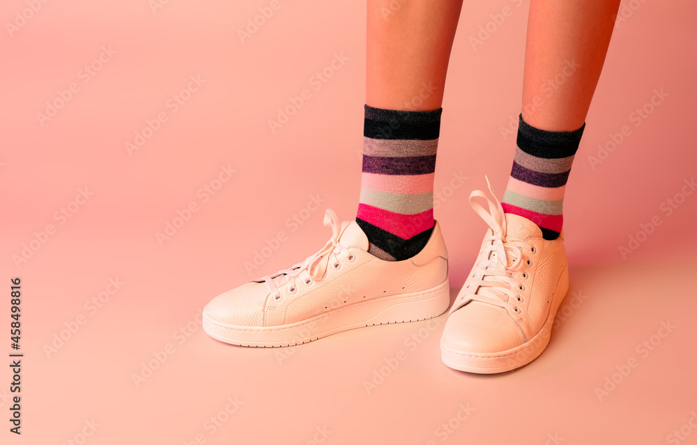 White leather sneakers shoes, colourful striped socks and girl’s legs on pastel pink