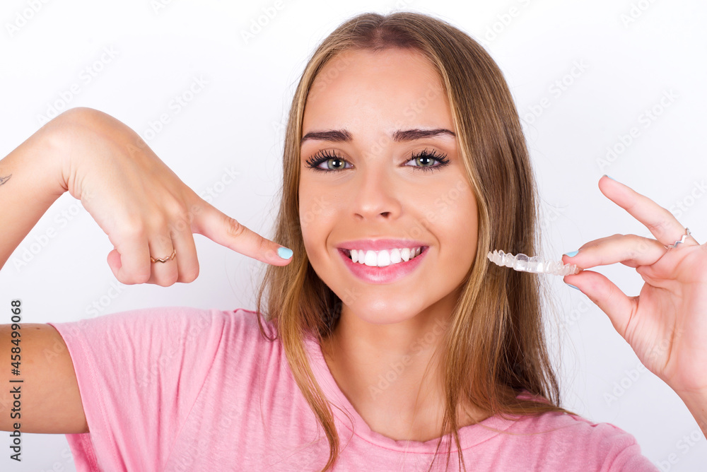 beautiful blonde girl wearing pink t-shirt on white background holding an invisible aligner and pointing to her perfect straight teeth. Dental healthcare and confidence concept. - obrazy, fototapety, plakaty 