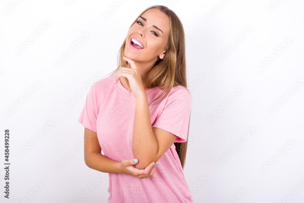 beautiful blonde girl wearing pink t-shirt on white background laughs happily keeps hand on chin expresses positive emotions smiles broadly has carefree expression beautiful blonde girl wearing pink t-shirt on white background laughs happily keeps hand on chin expresses positive emotions smiles broadly has carefree expression