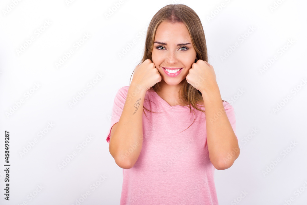 Happy beautiful blonde girl wearing pink t-shirt on white background keeps fists on cheeks smiles broadly and has positive expression being in good mood Happy beautiful blonde girl wearing pink t-shirt on white background keeps fists on cheeks smiles broadly and has positive expression being in good mood