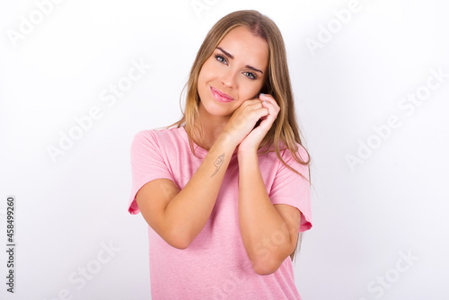Charming serious beautiful blonde girl wearing pink t-shirt on white background keeps hands near face smiles tenderly at camera
