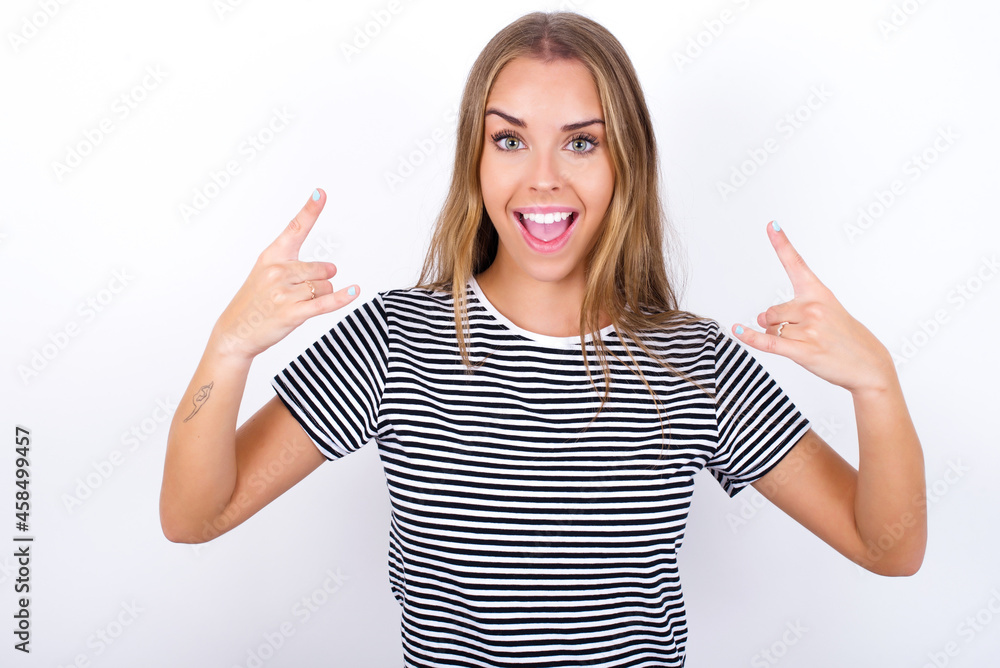 beautiful blonde girl wearing striped t-shirt on white background makes rock n roll sign looks self confident and cheerful enjoys cool music at party. Body language concept. beautiful blonde girl wearing striped t-shirt on white background makes rock n roll sign looks self confident and cheerful enjoys cool music at party. Body language concept.