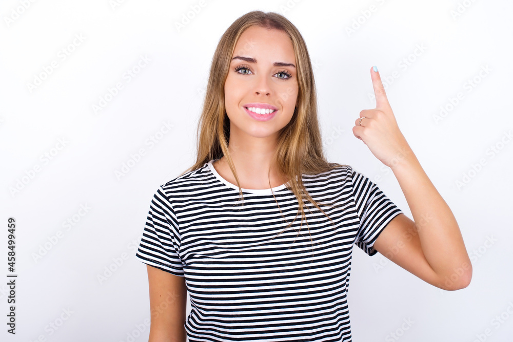 beautiful blonde girl wearing striped t-shirt on white background smiling and looking friendly, showing number one or first with hand forward, counting down beautiful blonde girl wearing striped t-shirt on white background smiling and looking friendly, showing number one or first with hand forward, counting down