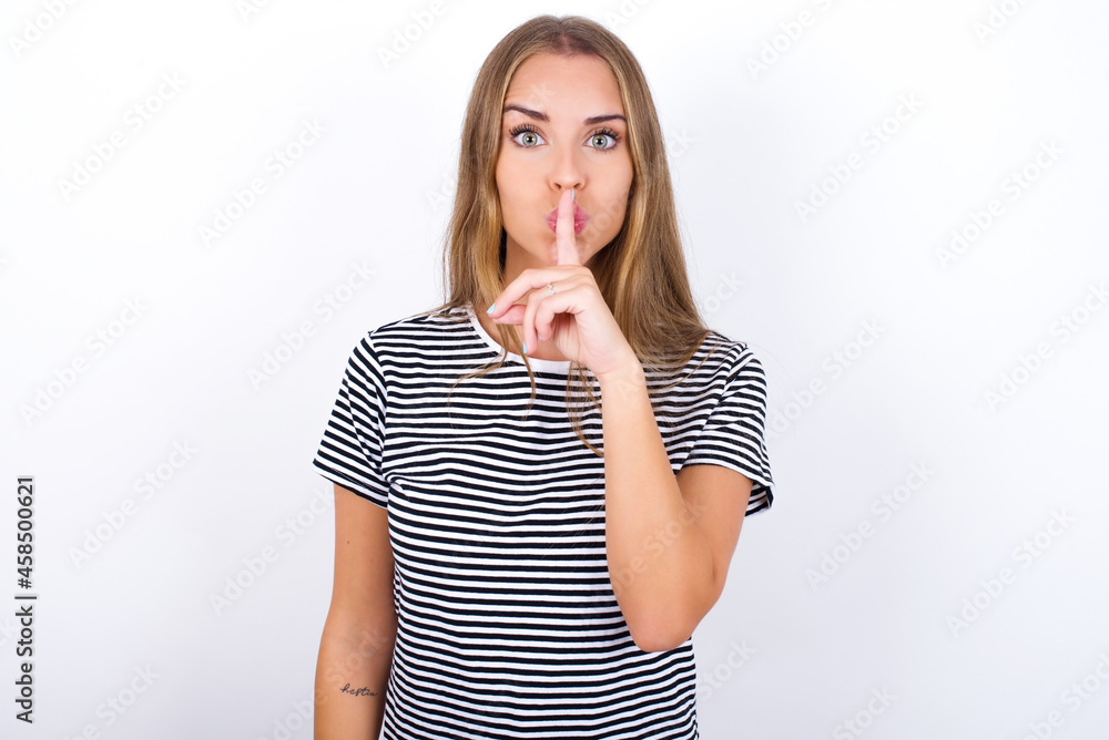 Surprised beautiful blonde girl wearing striped t-shirt on white background makes silence gesture, keeps finger over lips and looks mysterious at camera Surprised beautiful blonde girl wearing striped t-shirt on white background makes silence gesture, keeps finger over lips and looks mysterious at camera