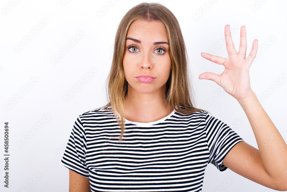 beautiful blonde girl wearing striped t-shirt on white background purses lip and gestures with hand, shows something very little. beautiful blonde girl wearing striped t-shirt on white background purses lip and gestures with hand, shows something very little.