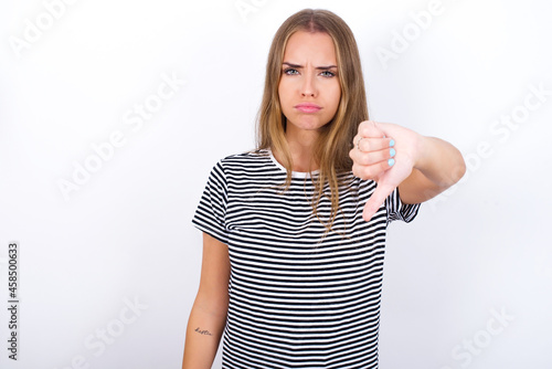 beautiful blonde girl wearing striped t-shirt on white background looking unhappy and angry showing rejection and negative with thumbs down gesture. Bad expression.