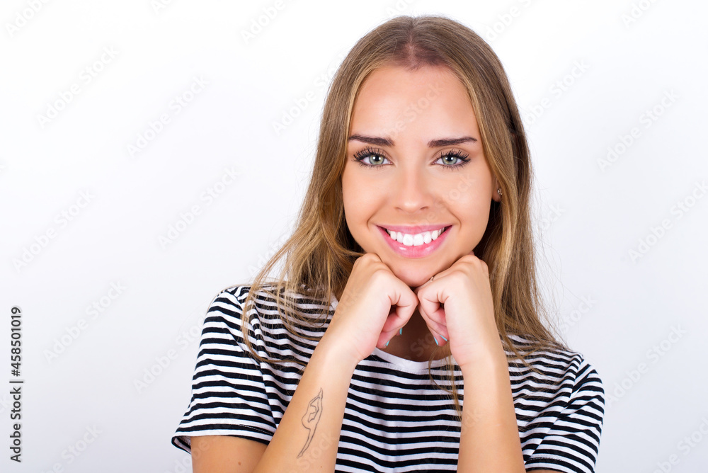 Satisfied beautiful blonde girl wearing striped t-shirt on white background touches chin with both hands, smiles pleasantly, rejoices good day with lover Satisfied beautiful blonde girl wearing striped t-shirt on white background touches chin with both hands, smiles pleasantly, rejoices good day with lover