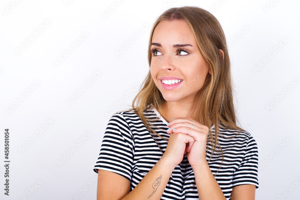 Happy beautiful blonde girl wearing striped t-shirt on white background anticipates something awesome happen, looks happily aside, keeps hands together near face, has glad expression. Happy beautiful blonde girl wearing striped t-shirt on white background anticipates something awesome happen, looks happily aside, keeps hands together near face, has glad expression.