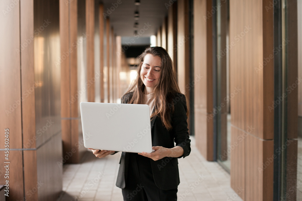 Young millennial businesswoman working on laptop near office building and smiling
