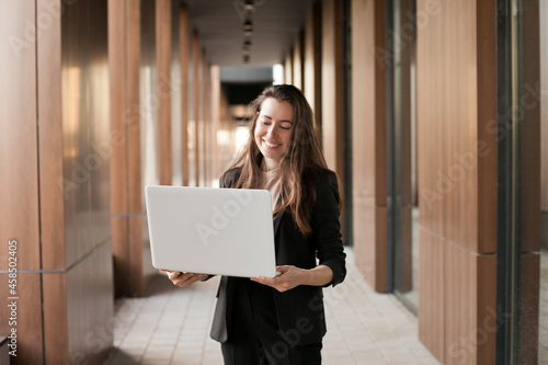 Young millennial businesswoman working on laptop near office building and smiling