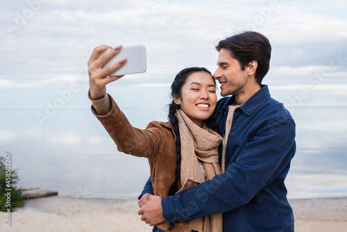Smiling man hugging asian girlfriend taking selfie on beach