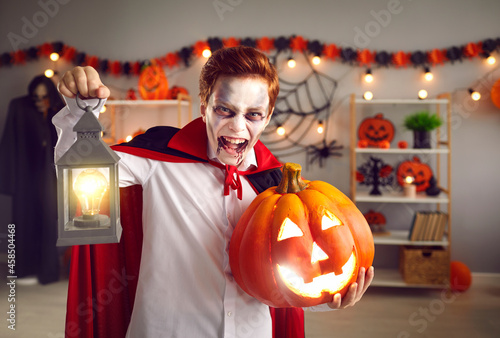 Trick or treat. Spooky little boy dressed in Halloween vampire costume screams and scares looking at camera. Child with scary makeup holds lantern and pumpkin in room with holiday decorations.