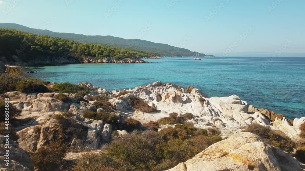 Aegean sea coast with greenery around, rocks, bushes and trees, blue water, moving yacht, Greece. Slow motion