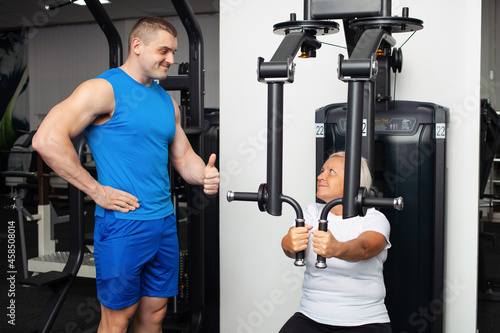 elderly woman is engaged in a simulator in the gym. The trainer helps and explains how to perform the exercise. healthy lifestyle, pensioner, concept, indoor. Sports training. thumbs up