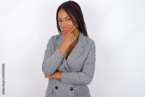 Thoughtful smiling Young African American businesswoman with braids on white wall keeps hand under chin, looks directly at camera, listens something with interest. Youth concept.