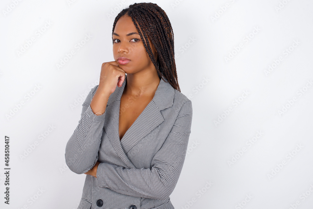 Portrait of thoughtful Young African American businesswoman with braids on white wall keeps hand under chin, looks away trying to remember something or listens something with interest. Youth concept. Portrait of thoughtful Young African American businesswoman with braids on white wall keeps hand under chin, looks away trying to remember something or listens something with interest. Youth concept.